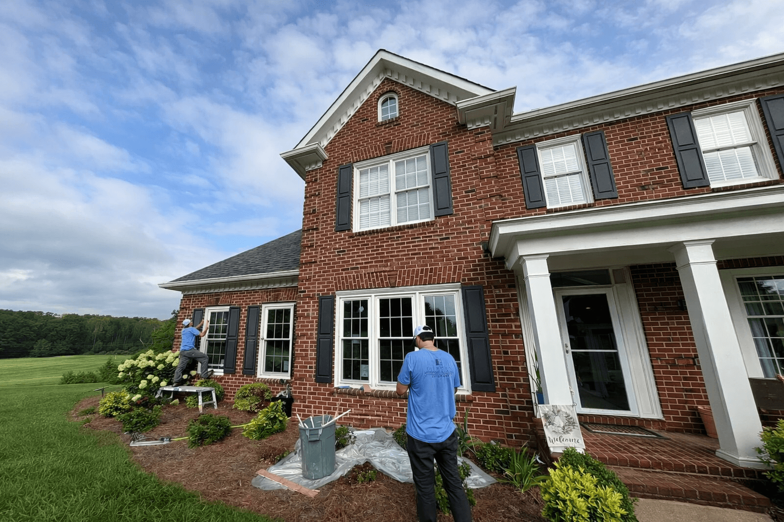 Two workers in blue shirts maintaining windows on a red brick house under a clear sky, demonstrating attention to detail and thorough service.